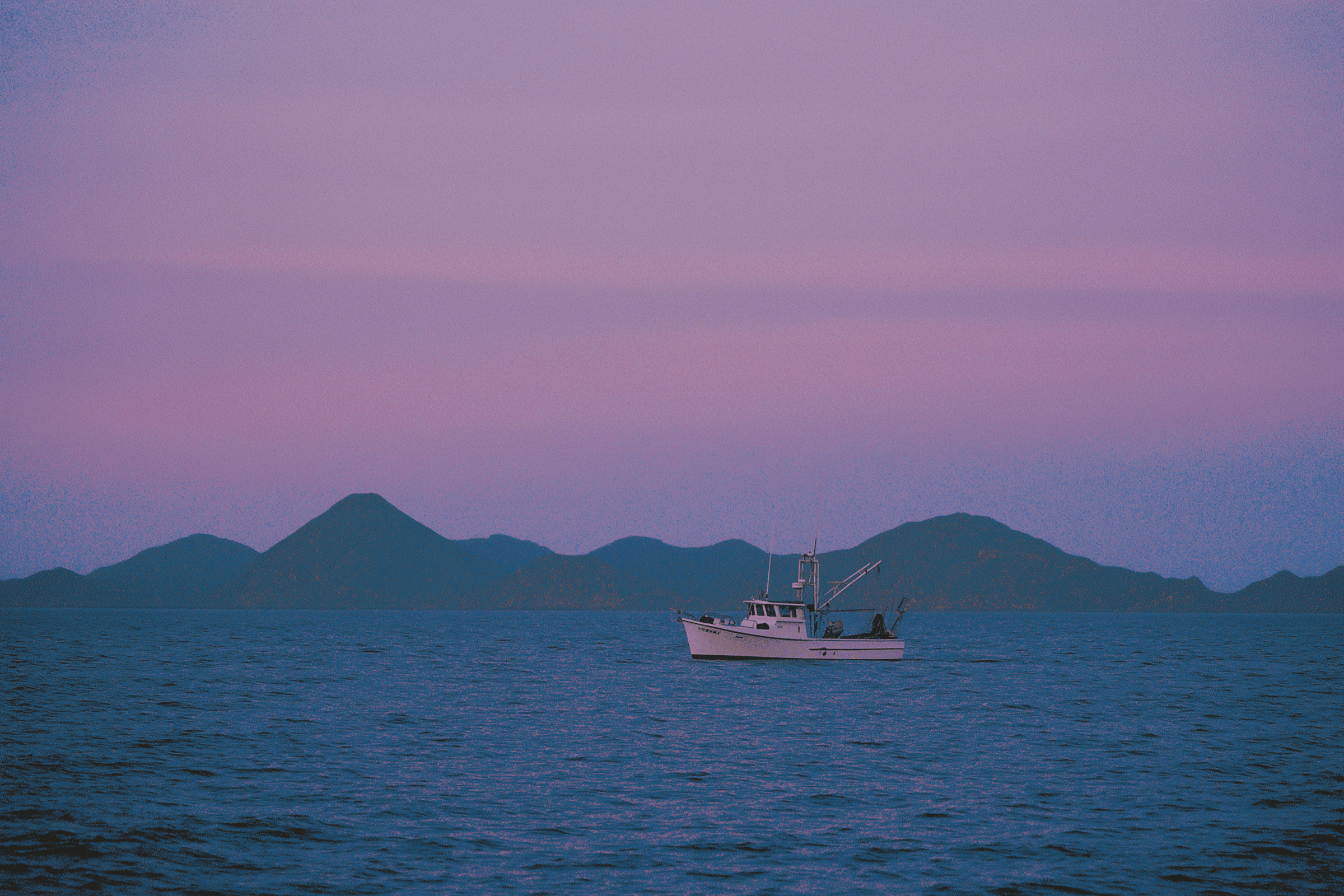 A lone fishing boat sails on the calm, deep blue ocean against a backdrop of rolling hills under a sky painted in hues of pink and purple during sunset.